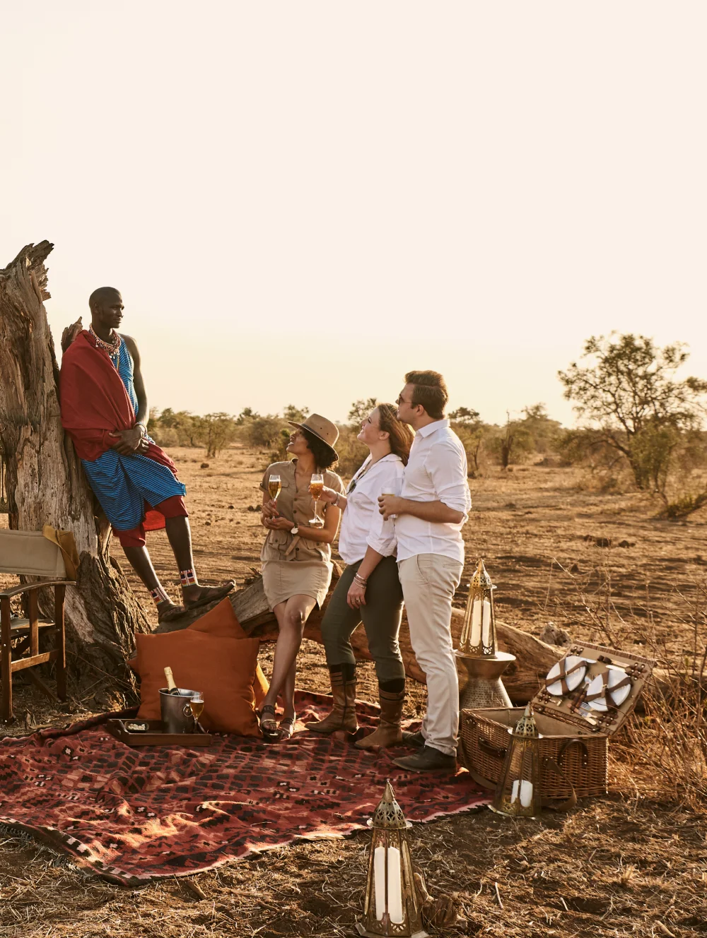 Maasai guide and guests with sundowners on a rug beside a fallen tree at sunset