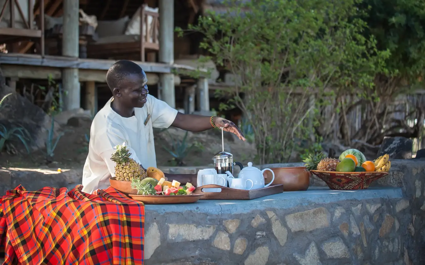 Staff member serving tea at outdoor dining table with fresh fruit, Apoka Safari Lodge, Kidepo Valley, Uganda