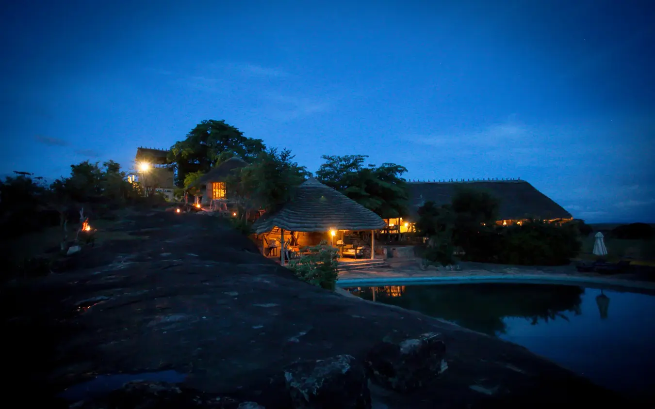 Illuminated lodge buildings at dusk beside water pool, Apoka Safari Lodge, Kidepo Valley, Uganda