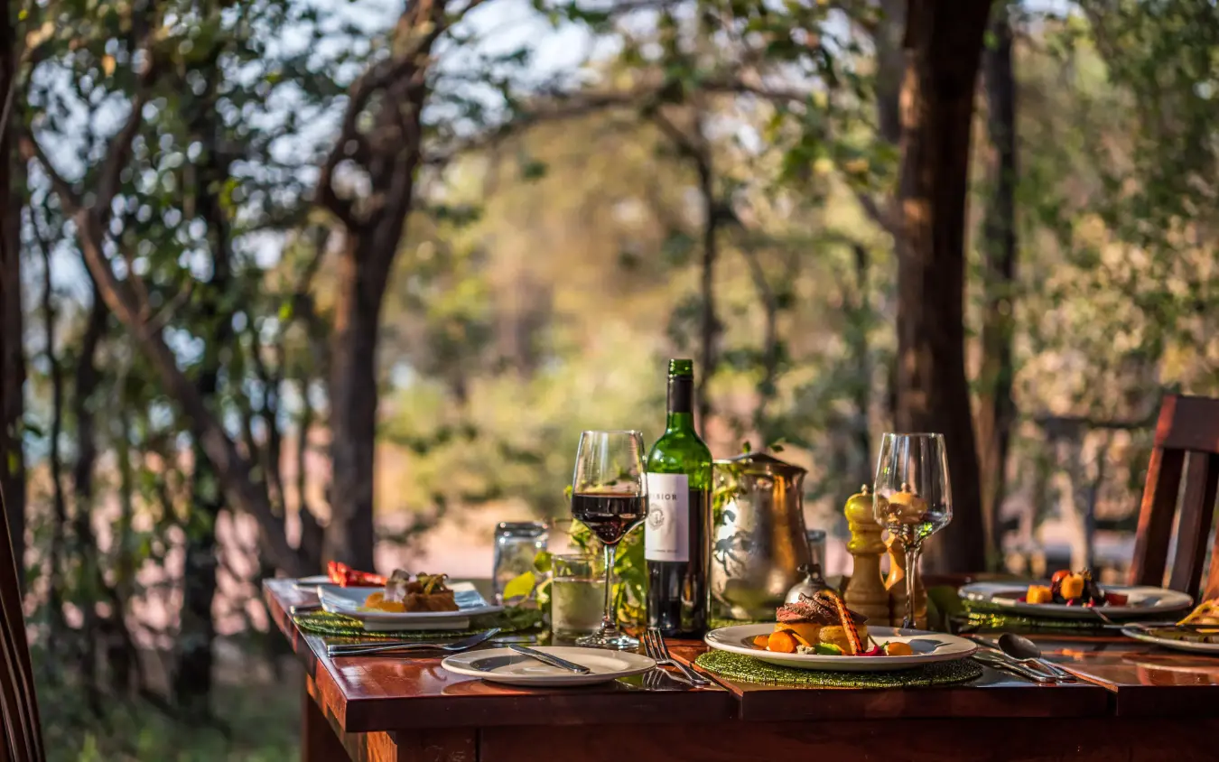 Outdoor dining table set among trees with wine and place settings, Changa Safari Camp, Matusadona National Park, Zimbabwe