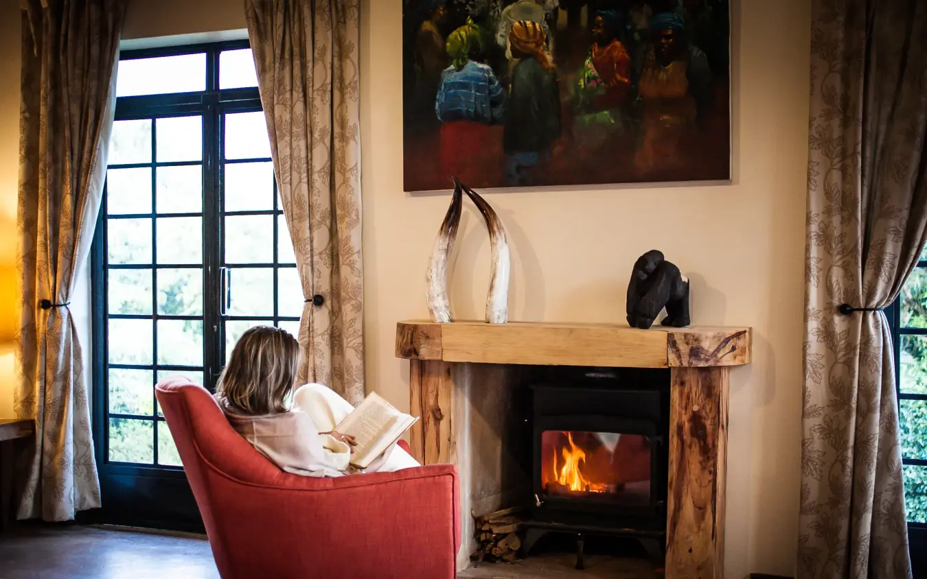 Woman reading by fireplace with mountain views through windows, Clouds Mountain Gorilla Lodge, Bwindi Impenetrable Forest, Uganda