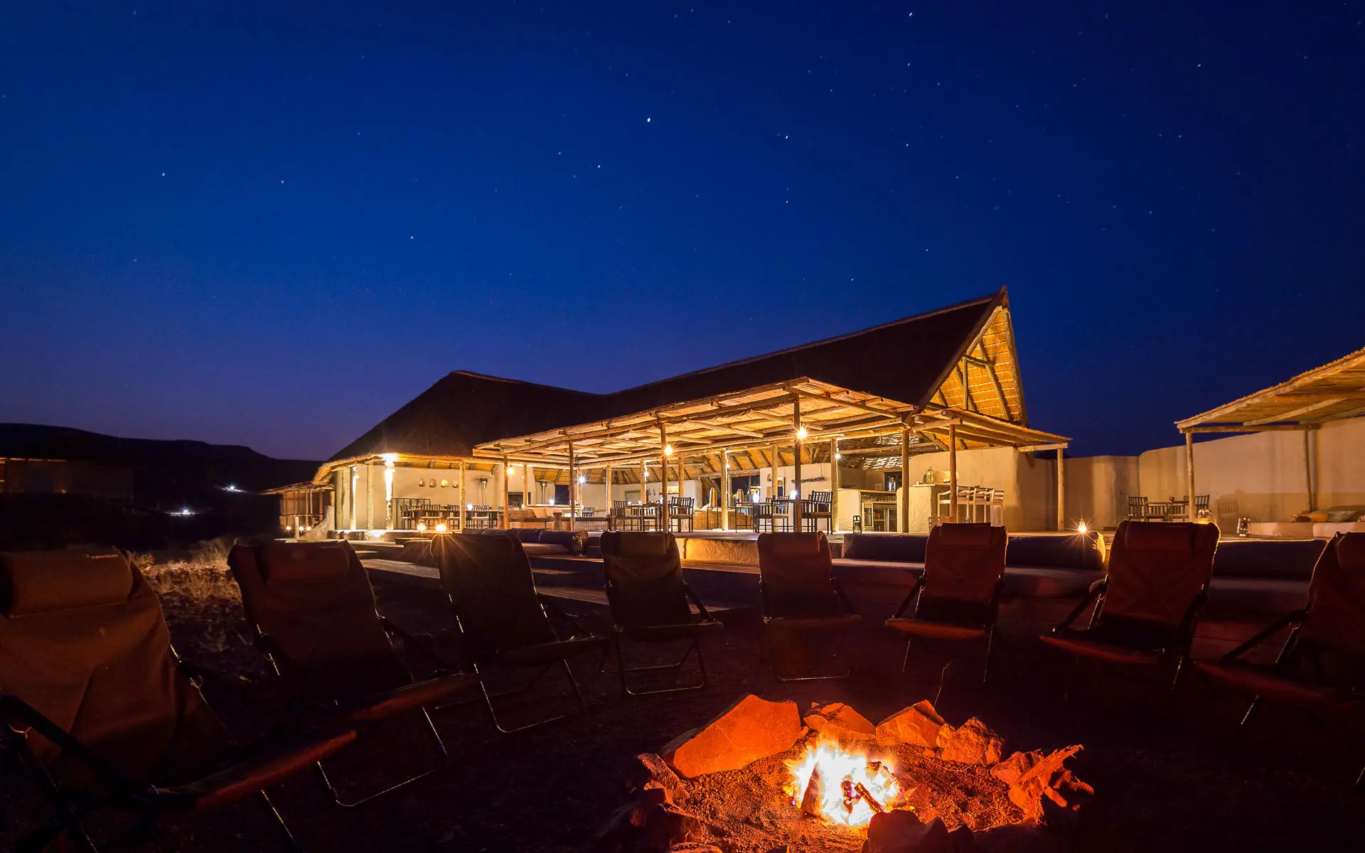 Illuminated main lodge at night with campfire in foreground, Damaraland Camp, Damaraland, Namibia