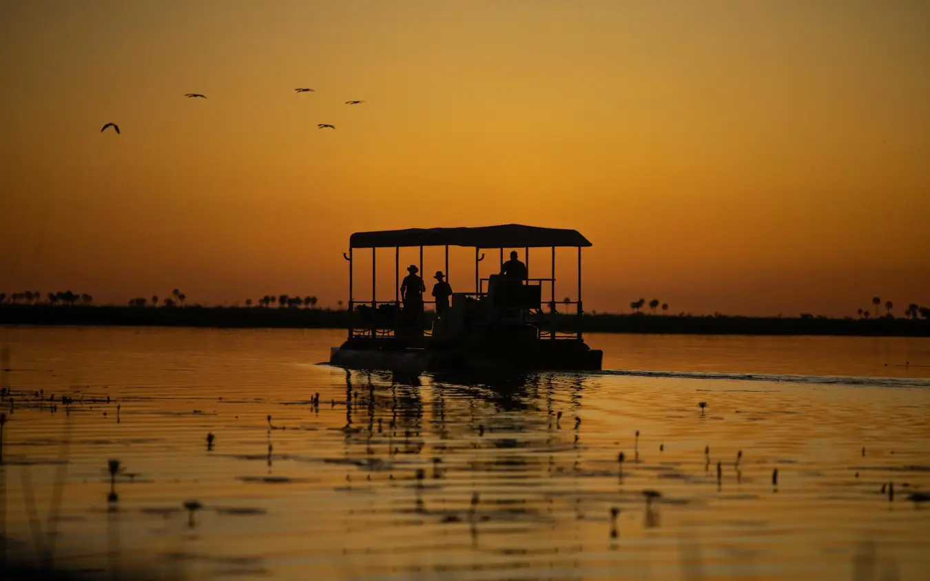 Silhouetted boat with passengers on calm water at sunset, DumaTau, Linyanti Wildlife Reserve, Botswana