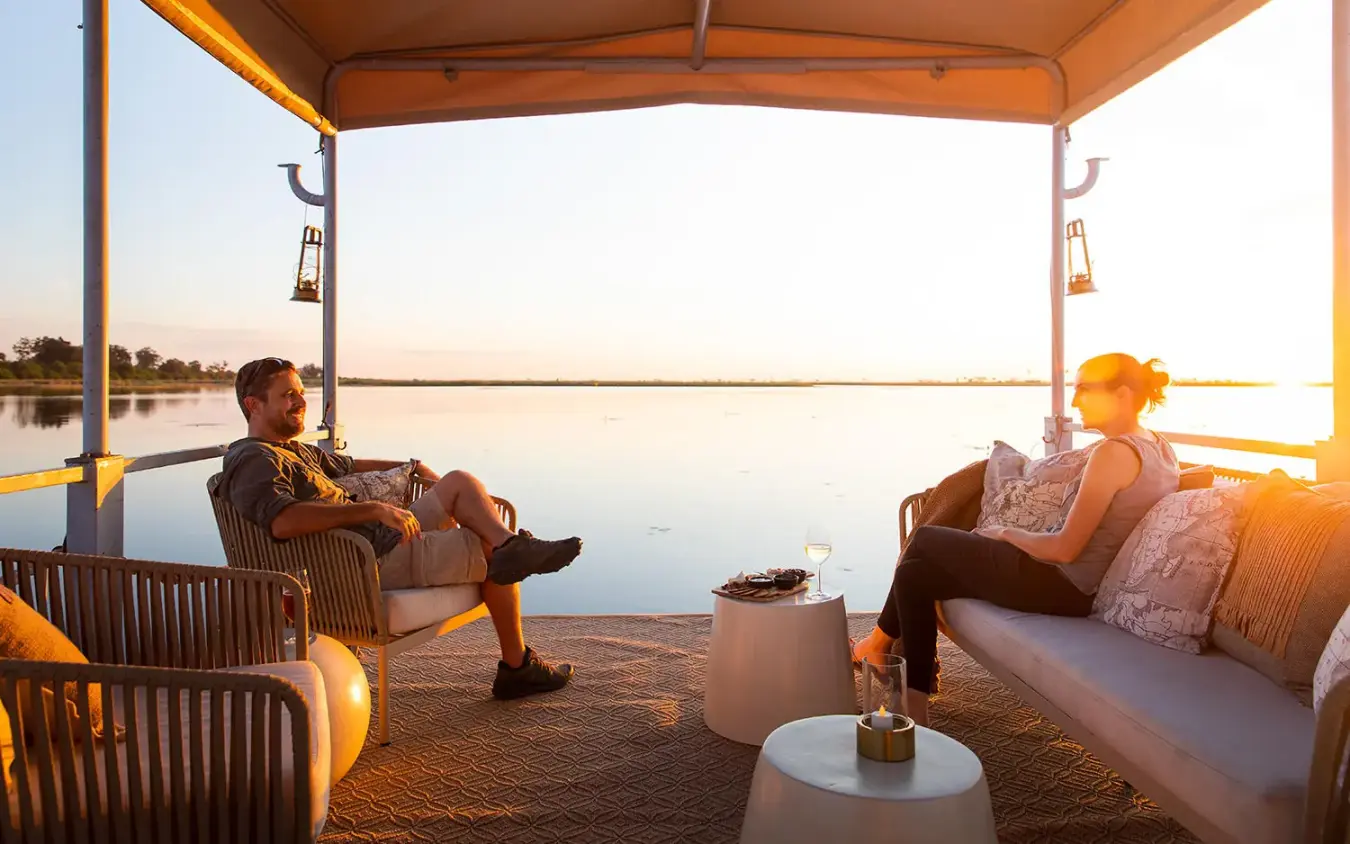 Couple relaxing on deck at sunset over water, DumaTau, Linyanti Wildlife Reserve, Botswana