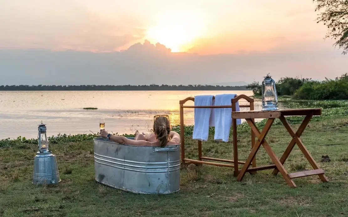 Person bathing in metal tub at sunset overlooking river, Honey Bear Bush Camp, Queen Elizabeth NP, Uganda