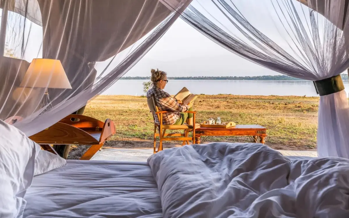 Guest reading in bed with lake view through sheer curtains, Honey Bear Bush Camp, Queen Elizabeth NP, Uganda