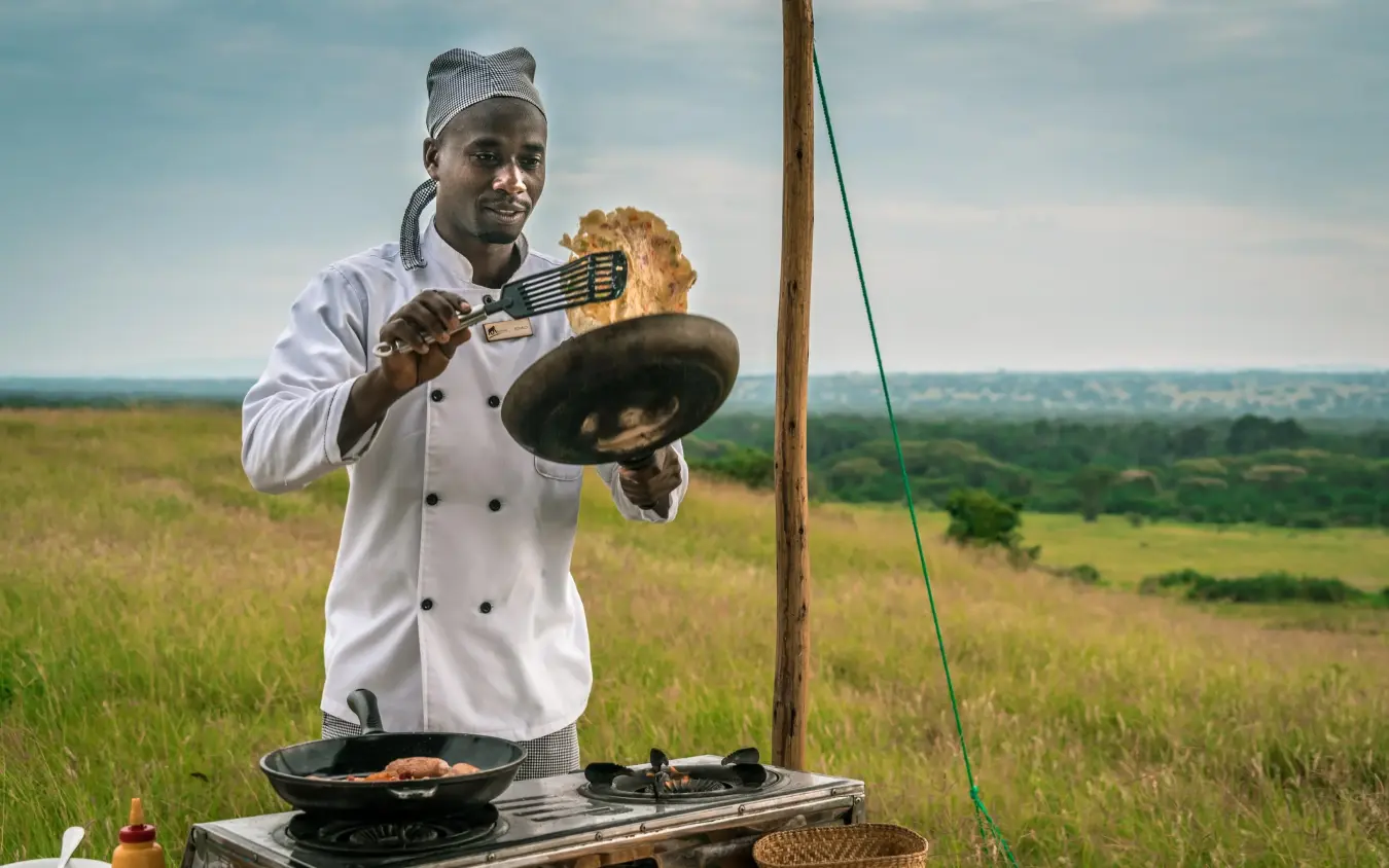 Chef cooking outdoors over portable stove in savanna landscape, Ishasha Wilderness Camp, Queen Elizabeth NP, Uganda