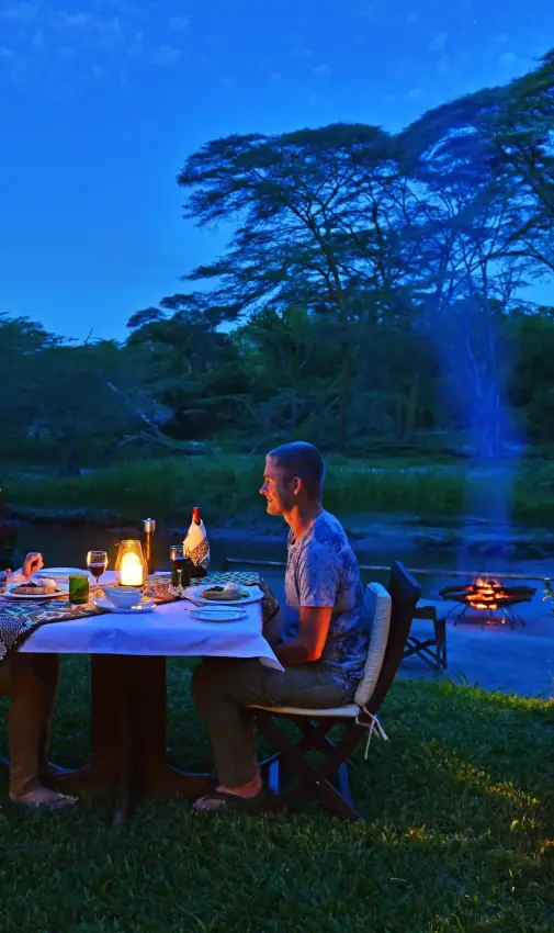 Couple dining by riverside at sunset with lantern lighting, Ishasha Wilderness Camp, Queen Elizabeth NP, Uganda
