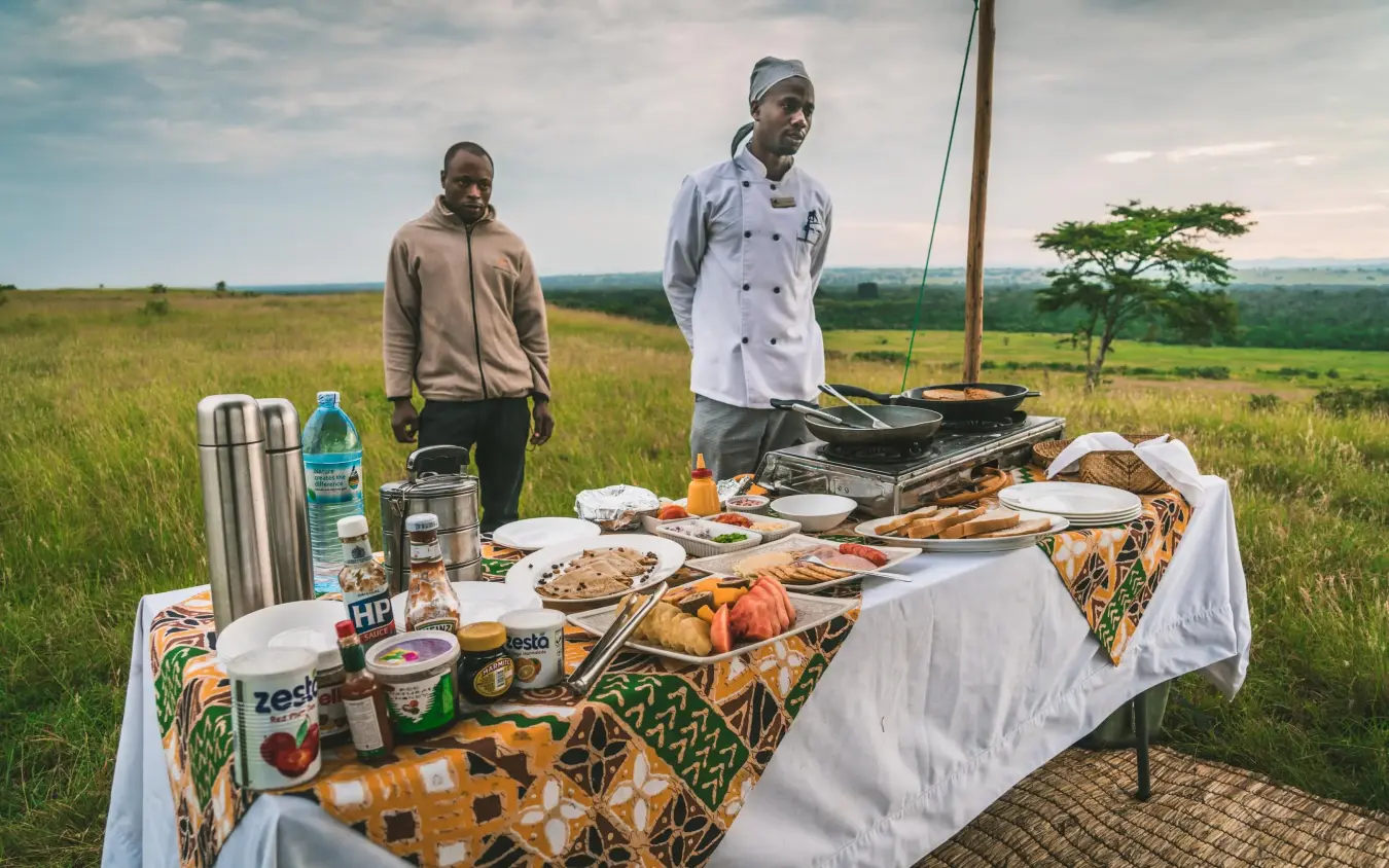 Outdoor dining setup with food and staff in grassland, Ishasha Wilderness Camp, Queen Elizabeth NP, Uganda