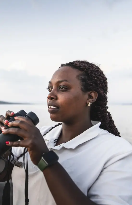 Woman with binoculars observing wildlife by calm water, Magashi Camp, Akagera National Park, Rwanda