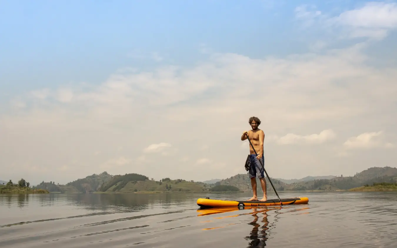 Guest stand-up paddleboarding on calm lake with hills, Mutanda Lake Resort, Lake Mutanda, Uganda