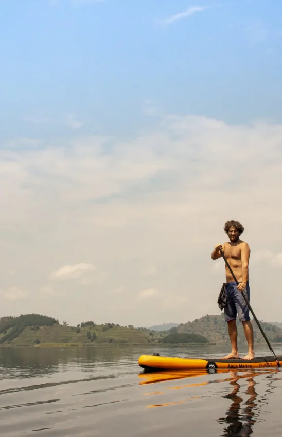 Guest stand-up paddleboarding on calm lake with hills, Mutanda Lake Resort, Lake Mutanda, Uganda