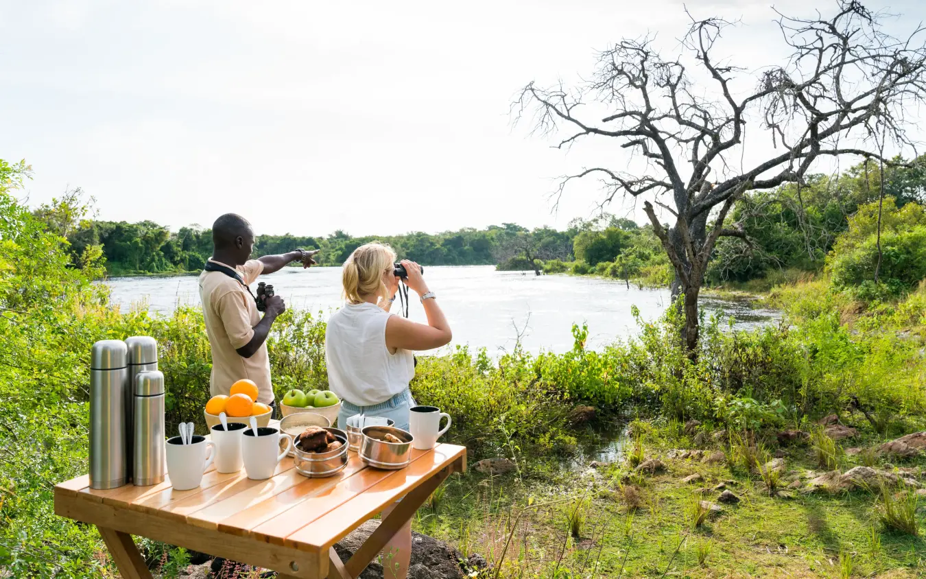 Guests observe wildlife with binoculars at riverside picnic setup, Papa’s Camp, Murchison Falls NP, Uganda