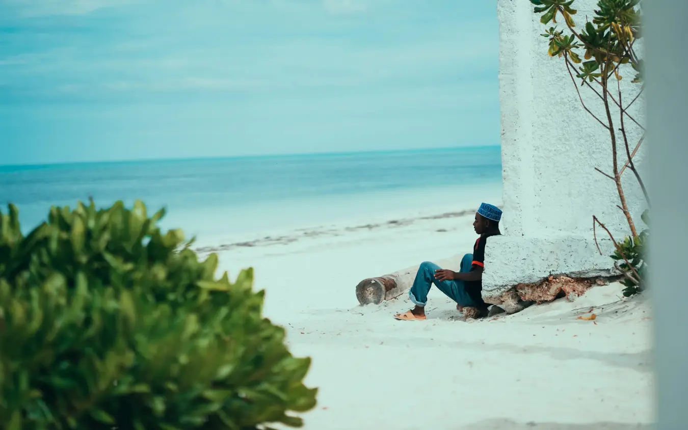Man in kufi cap sitting alone on white sand beach by wall