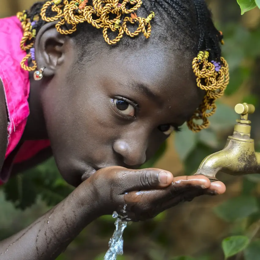 Child drinking water from outdoor tap with green foliage