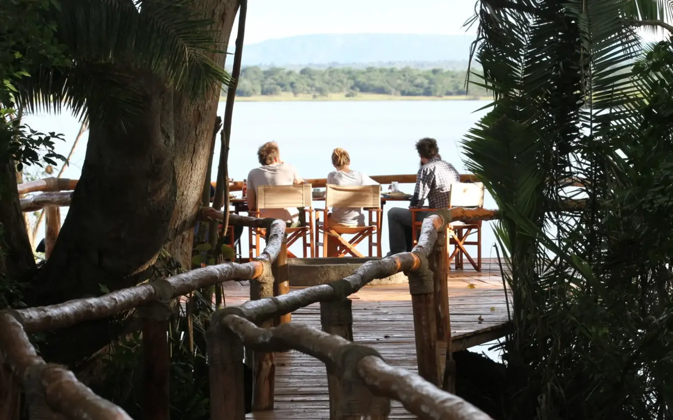 Guests relax on wooden deck overlooking river and forest, Ruzizi Tented Lodge, Akagera National Park, Rwanda
