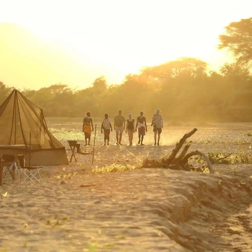 Group of people standing on riverbank at sunrise with tents, Sarara Treehouses, Northern Kenya, Kenya