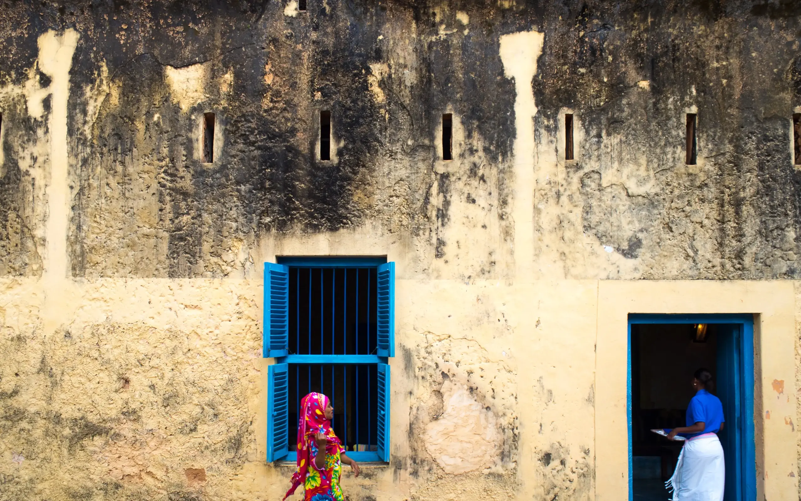 Woman in colorful sari walks past aged Zanzibar Stone Town building with blue shutters
