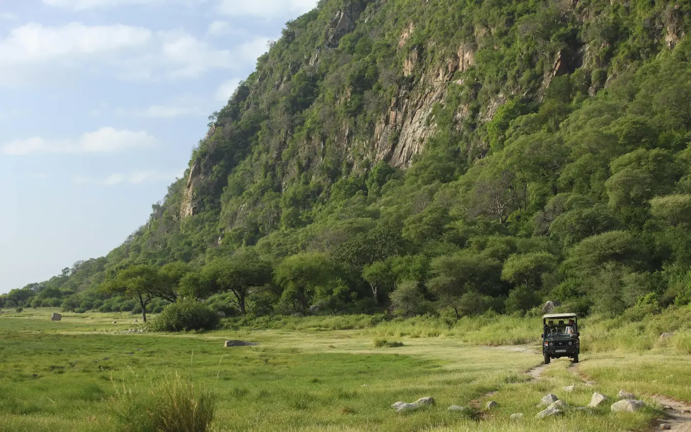 Safari vehicle on grassy plain beneath dramatic cliff face, Lake Manyara Tree Lodge, Lake Manyara, Tanzania