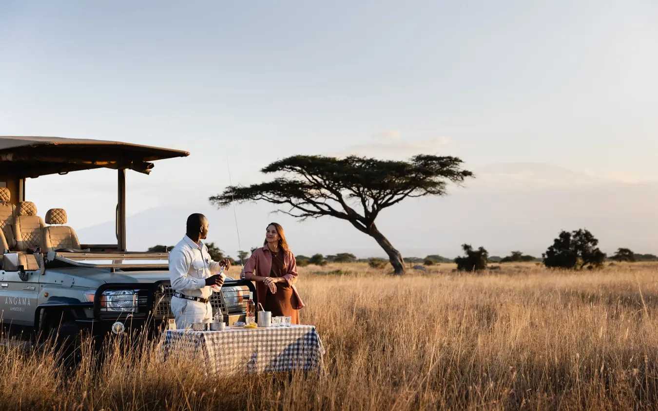 Couple enjoying sunset cocktails at safari vehicle in grassland, Angama Amboseli, Amboseli and Chyulu Hills, Kenya