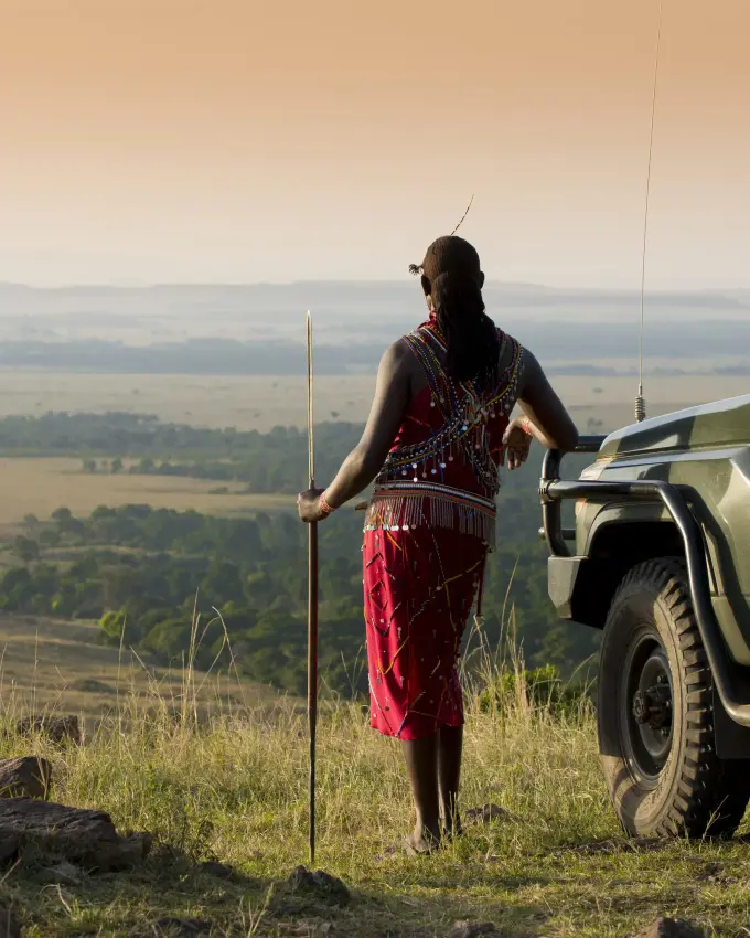 Guest in traditional dress standing beside safari vehicle overlooking valley, Maasai Mara, Kenya