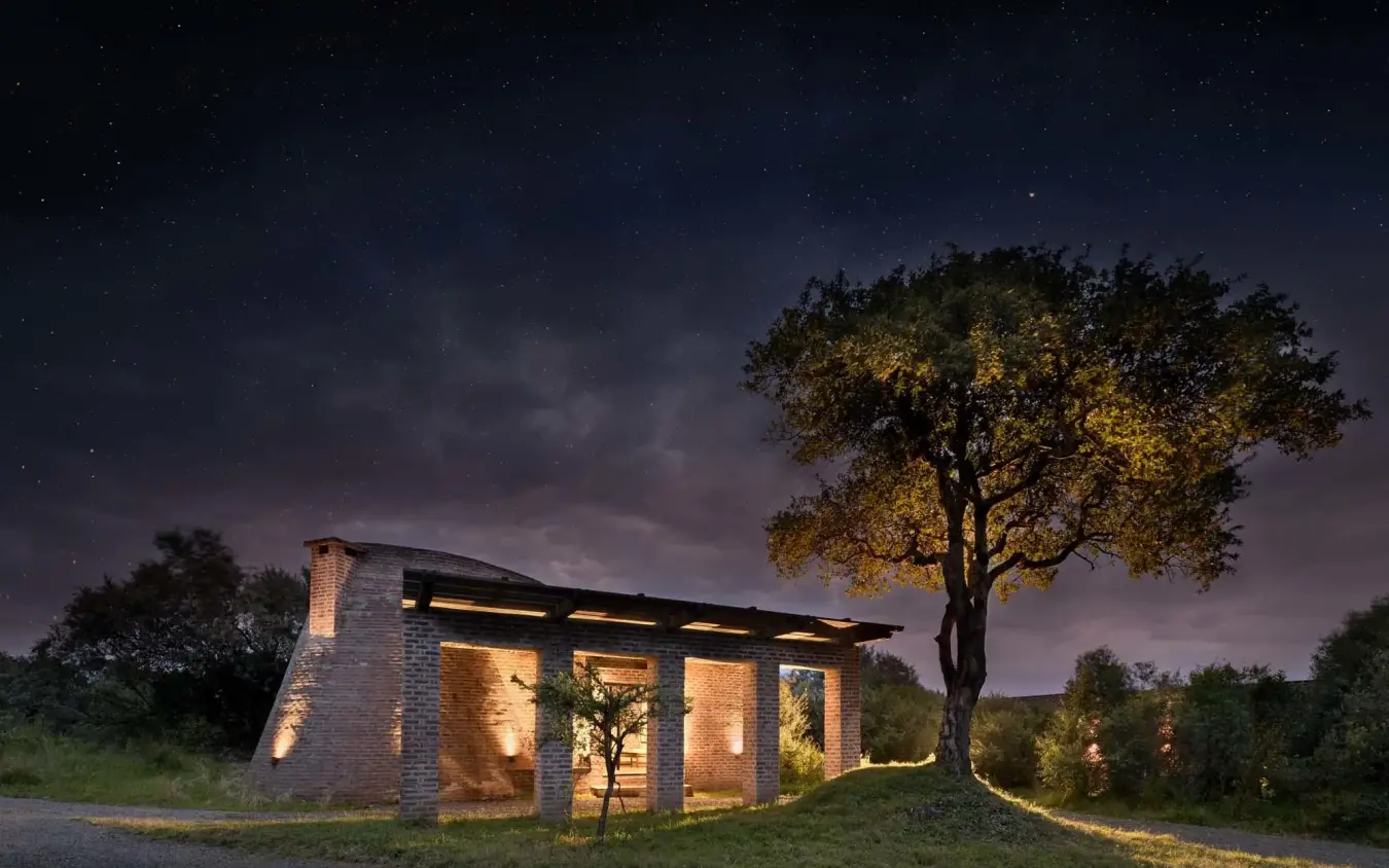 Modern brick lodge illuminated at dusk beneath acacia tree, Angama Mara, Maasai Mara, Kenya