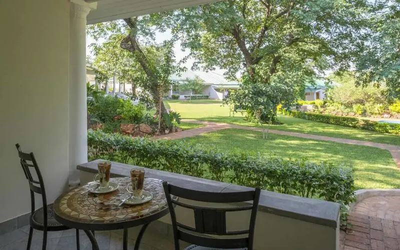 Covered porch with round table overlooking manicured gardens and grounds, Batonka Guest Lodge, Victoria Falls, Zimbabwe