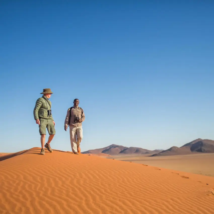 Two guests walking across red sand dunes with mountains, Sossusvlei, Namibia