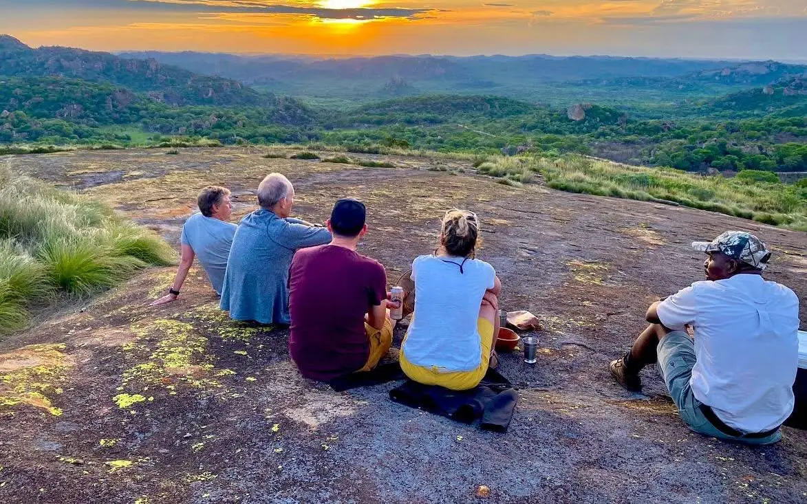 Group of hikers sitting on rocky plateau overlooking forested valley at sunset, Big Cave Camp, Matobo, Zimbabwe
