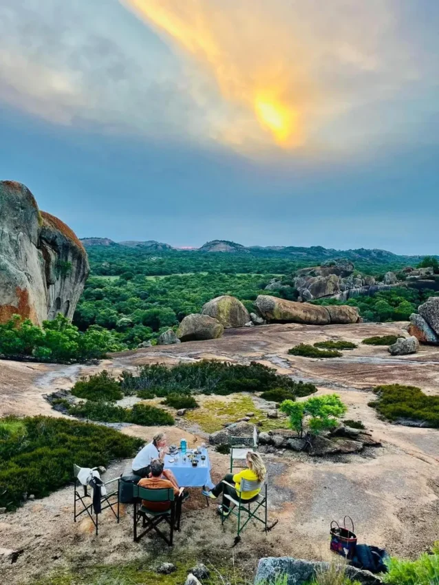 Couple dining at table on rocky plateau with granite boulders and vegetation, Big Cave Camp, Matobo, Zimbabwe