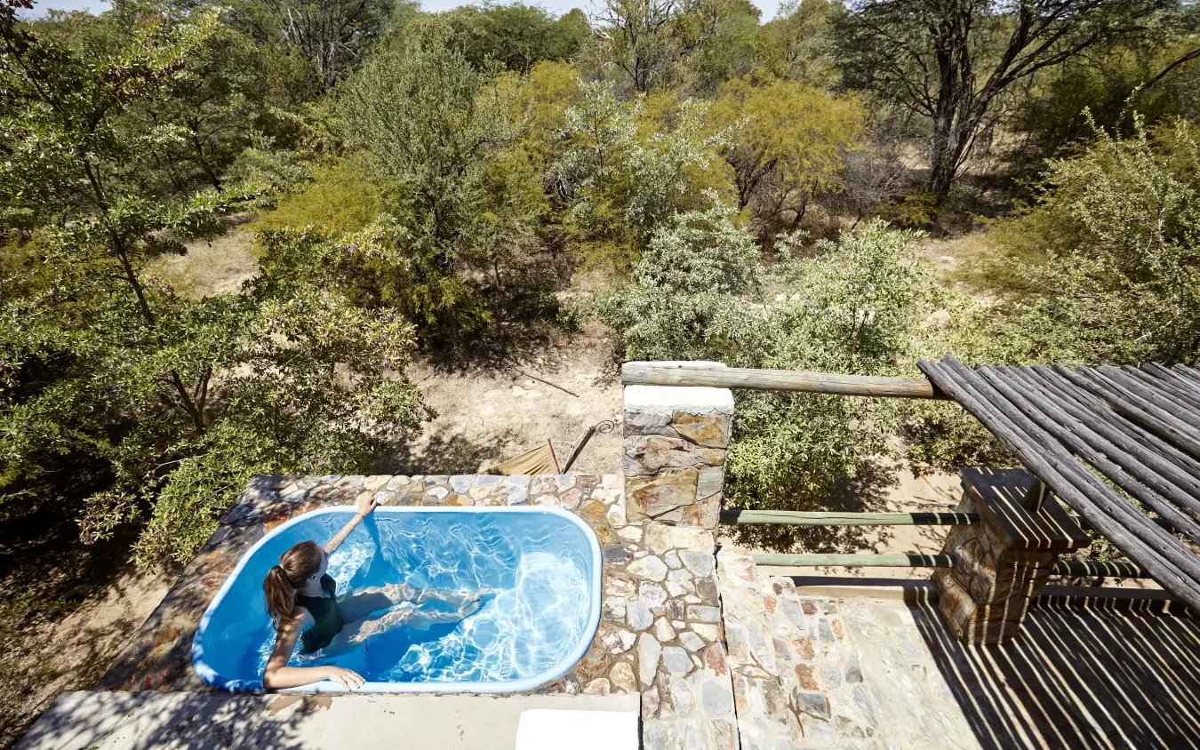 Guest relaxing in blue plunge pool overlooking wooded savanna landscape, Camelthorn Lodge, Hwange National Park, Zimbabwe
