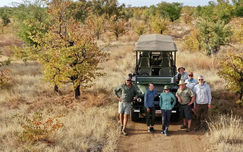 Safari vehicle with group of guests posing on dirt road in woodland savanna, Camp Chitubu, Hwange National Park, Zimbabwe