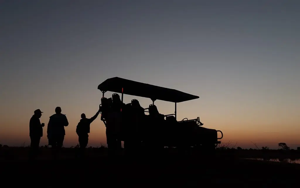 Safari vehicle with guests at dawn beside water in grassland, Camp Chitubu, Hwange National Park, Zimbabwe