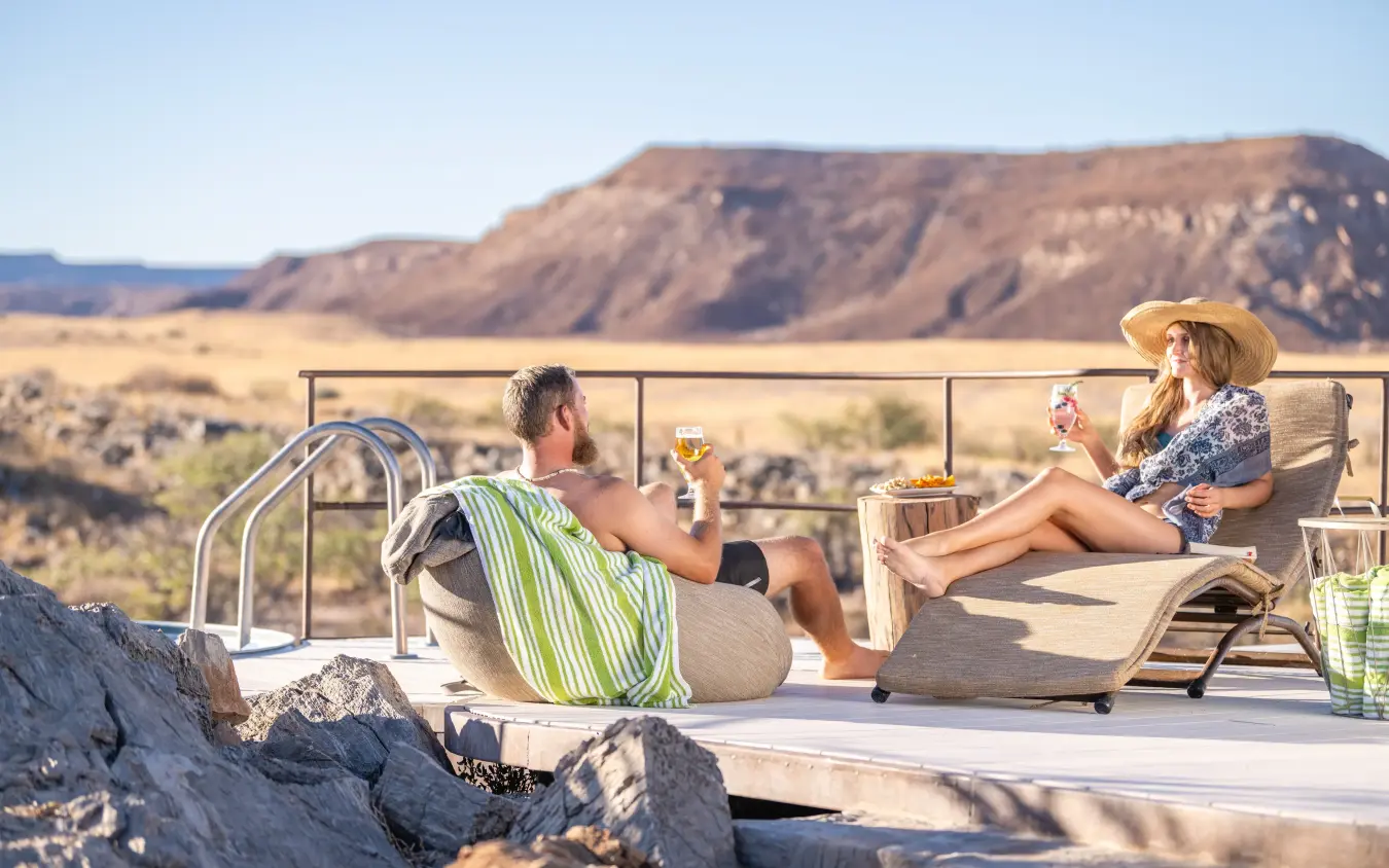 Couple relaxing on deck with drinks overlooking arid desert landscape, Camp Doros, Damaraland, Namibia