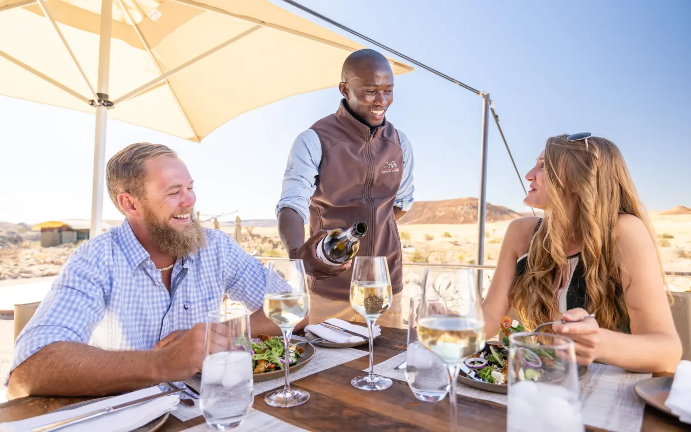 Server pouring wine at outdoor dining table under parasol in arid landscape, Camp Doros, Damaraland, Namibia