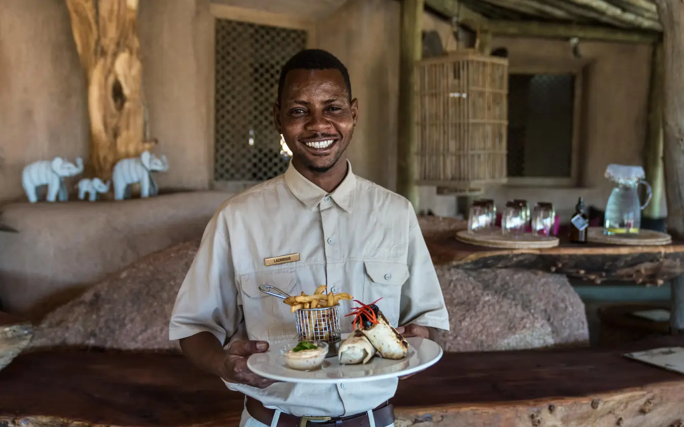 Staff member presenting plated food in rustic lodge interior, Camp Kipwe, Damaraland, Namibia