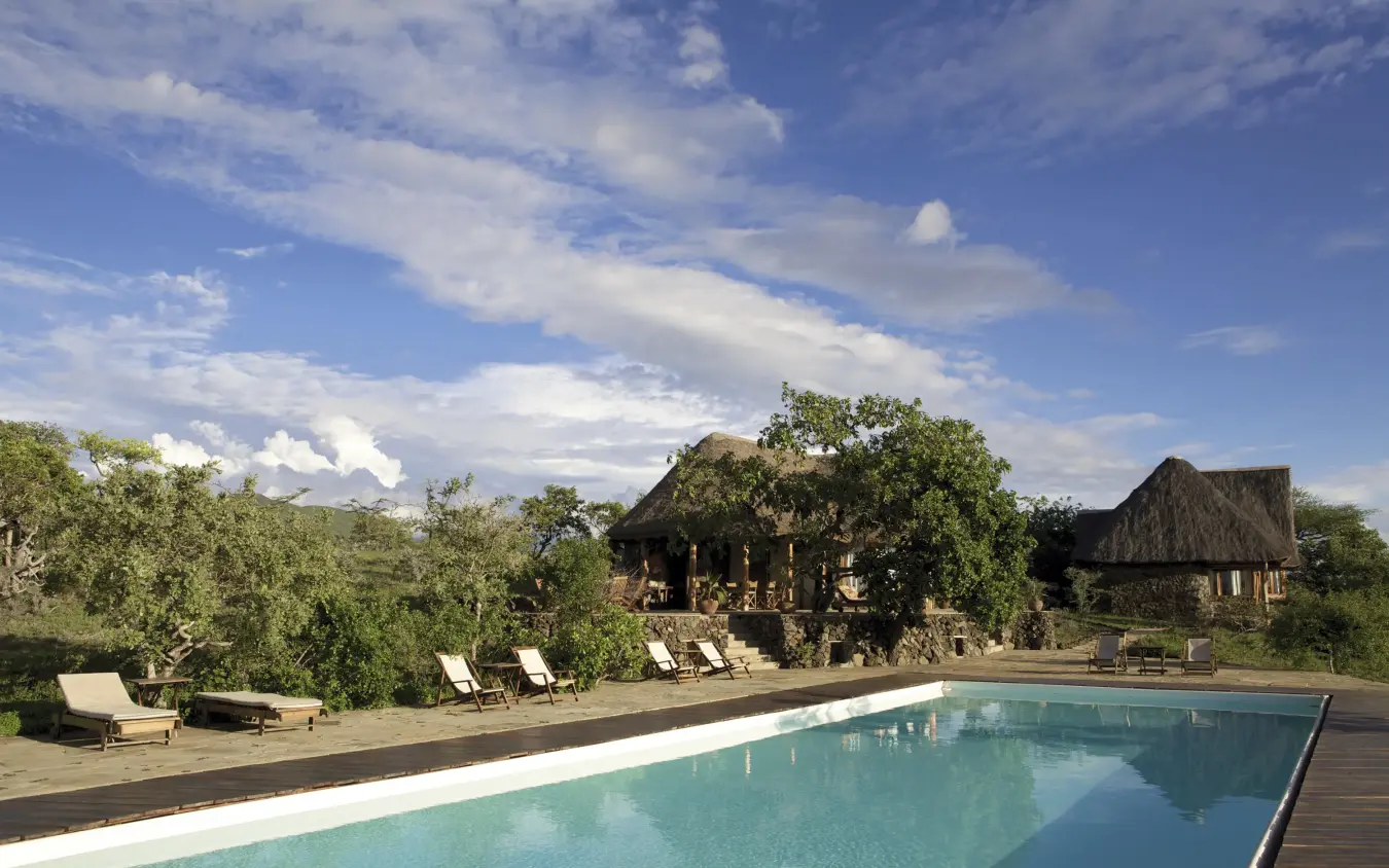 Rectangular swimming pool with thatched-roof lodges and savanna landscape, Campi ya Kanzi, Amboseli and Chyulu Hills, Kenya