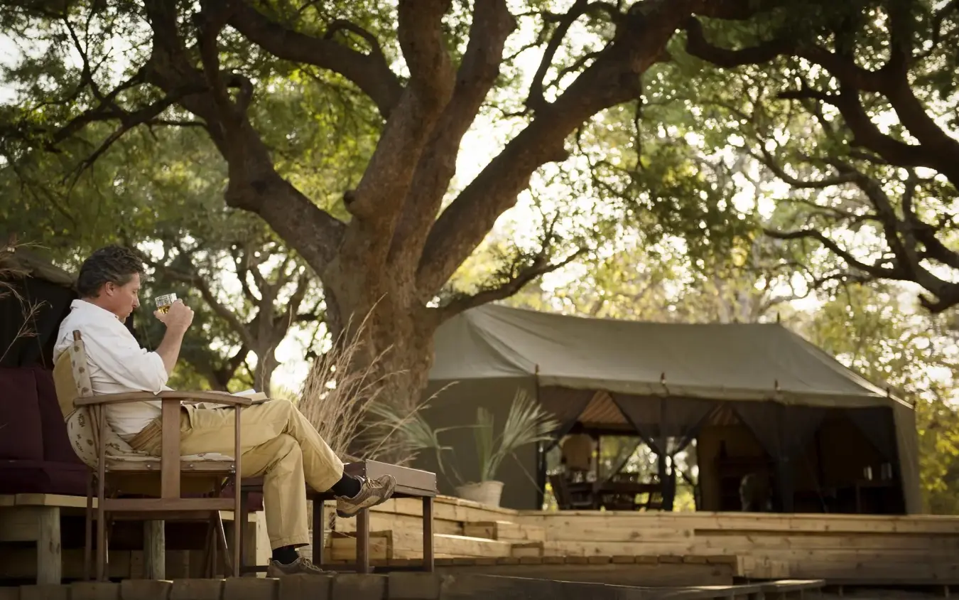 Guest relaxing on wooden deck beneath large trees, Chada Camp, Katavi National Park, Tanzania
