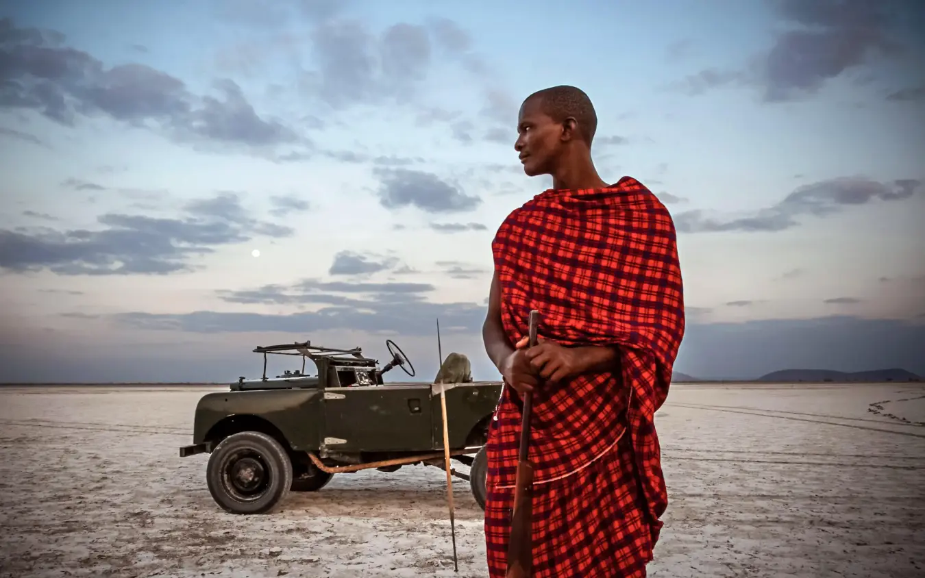 Maasai guide standing by vintage safari vehicle at sunset, Chem Chem Lodge, Lake Manyara, Tanzania