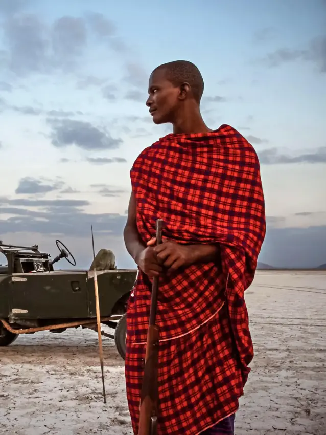 Maasai guide standing by vintage safari vehicle at sunset, Chem Chem Lodge, Lake Manyara, Tanzania
