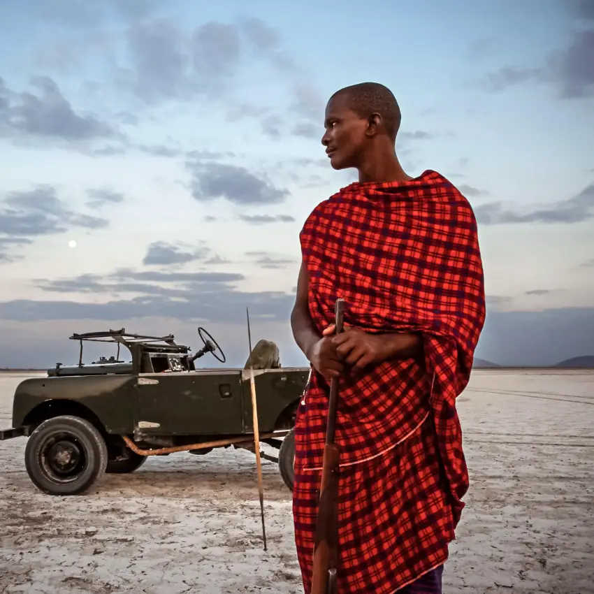 Maasai guide standing by vintage safari vehicle at sunset, Chem Chem Lodge, Lake Manyara, Tanzania