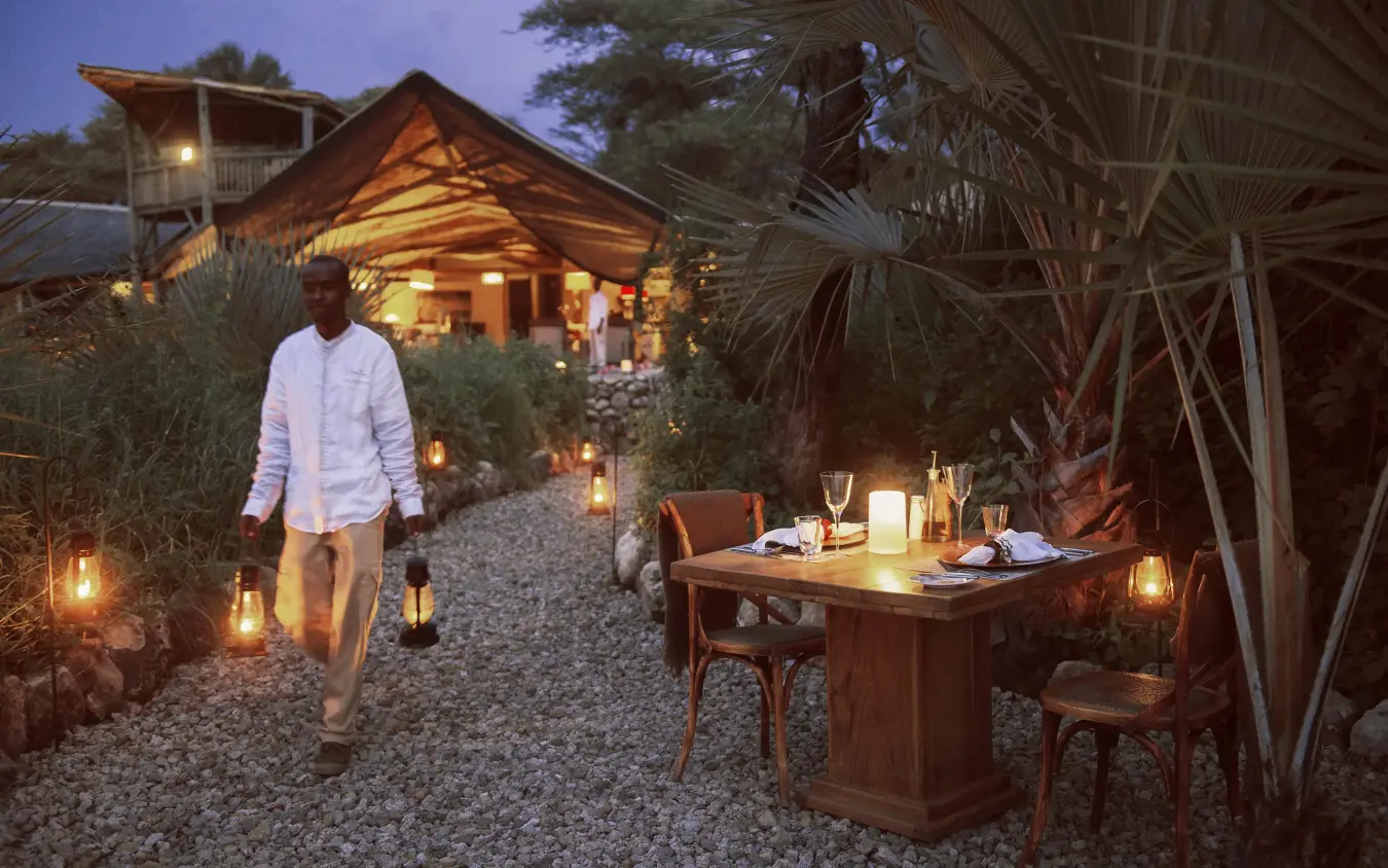 Guest walking toward illuminated safari lodge at dusk with dining setup, Chem Chem Lodge, Lake Manyara, Tanzania