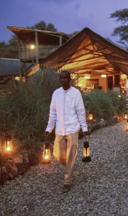 Guest walking toward illuminated safari lodge at dusk with dining setup, Chem Chem Lodge, Lake Manyara, Tanzania