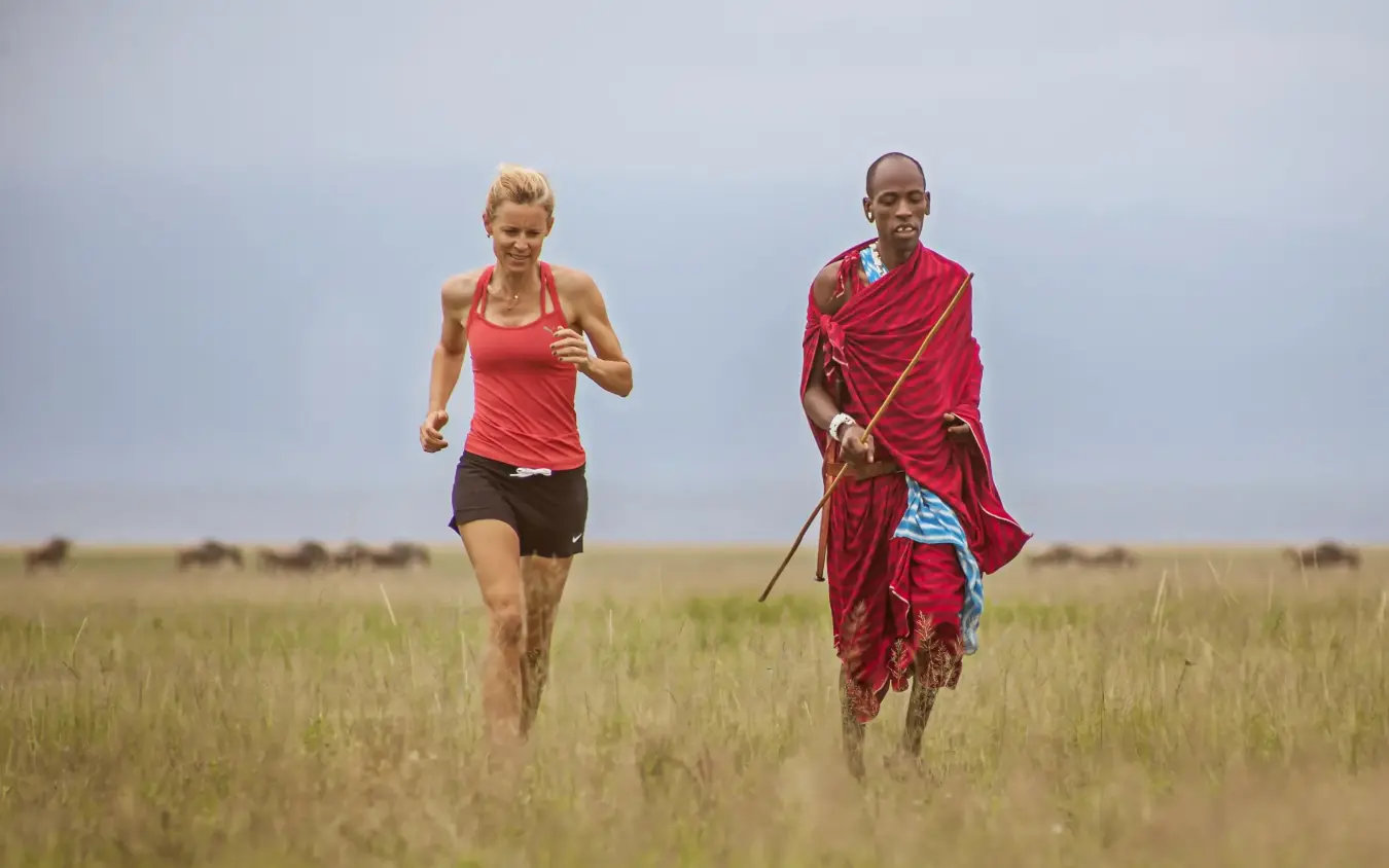 Woman and Maasai warrior jogging together across grassland savanna, Chem Chem Lodge, Lake Manyara, Tanzania