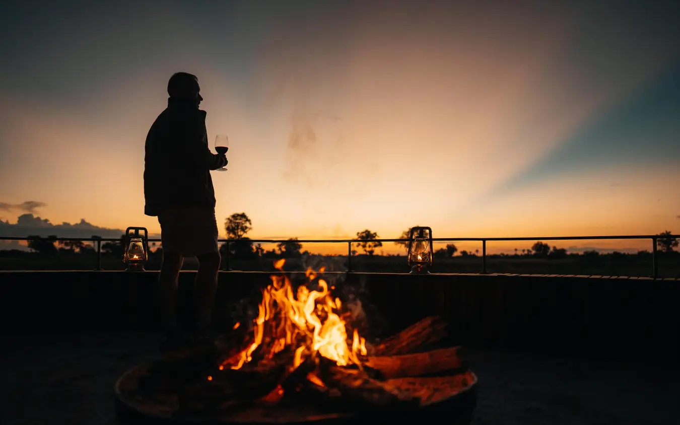 Person standing by campfire at sunset with bridge, Chitabe Camp, The Okavango Delta, Botswana