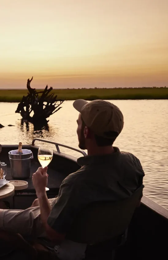 Man with wine glass on boat at sunset overlooking wetland, Chobe Chilwero, Chobe, Botswana
