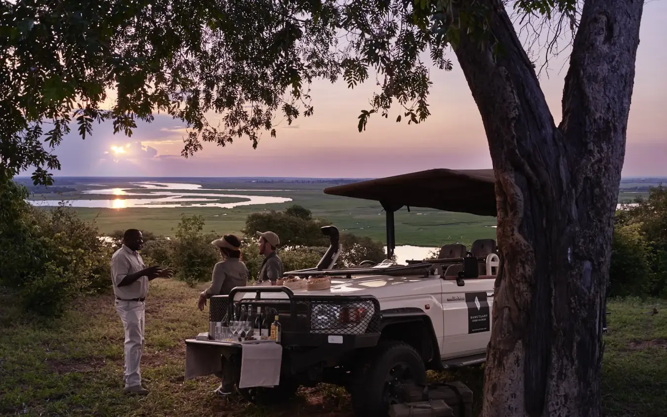 Safari vehicle parked under tree at sunset overlooking river valley, Chobe Chilwero, Chobe, Botswana