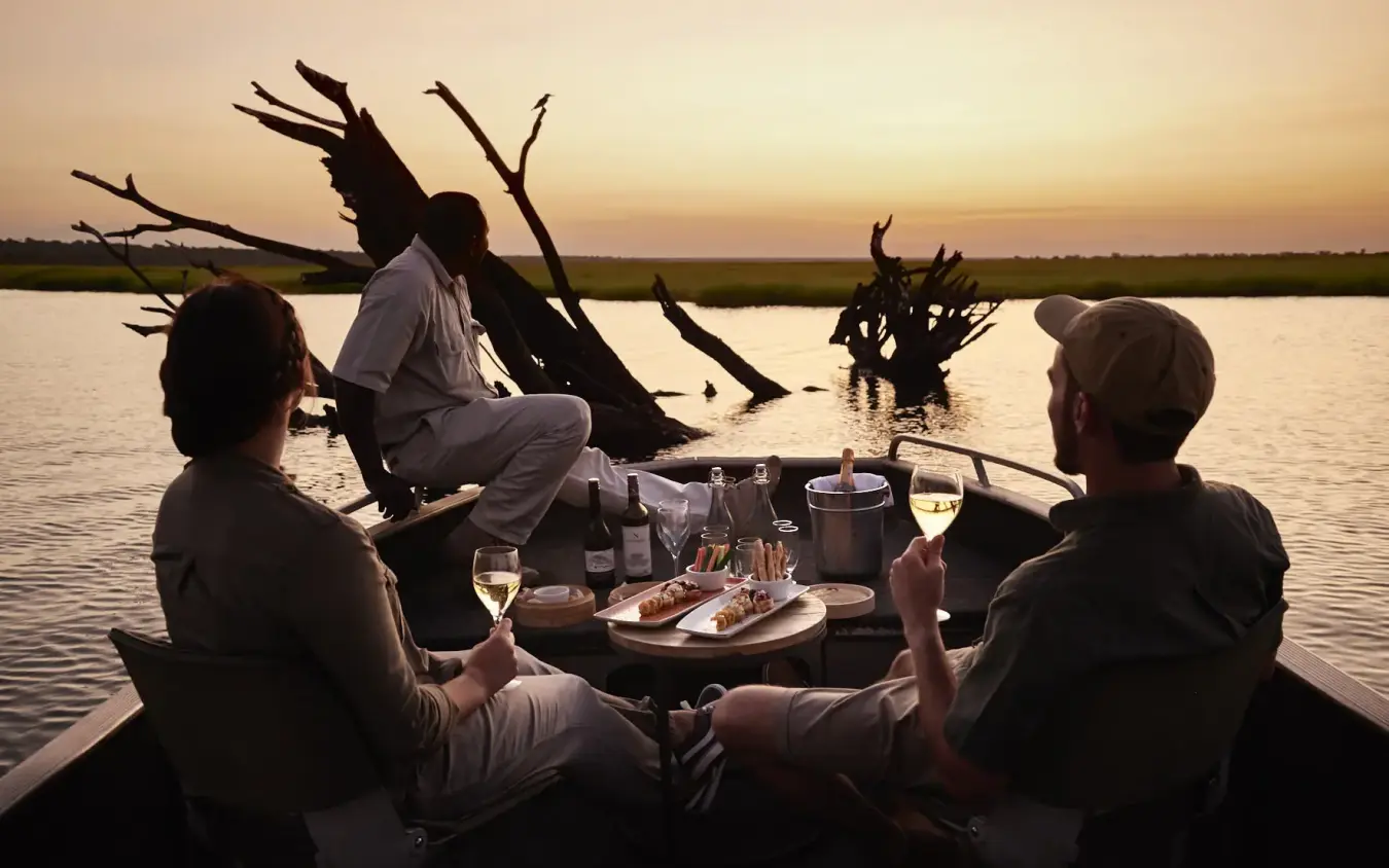 Guests dining on boat at sunset with dead trees in waterway, Chobe Chilwero, Chobe, Botswana