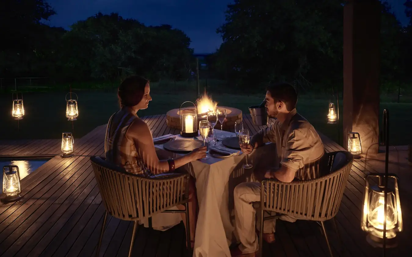 Couple dining at candlelit table on wooden deck at dusk, Chobe Chilwero, Chobe, Botswana
