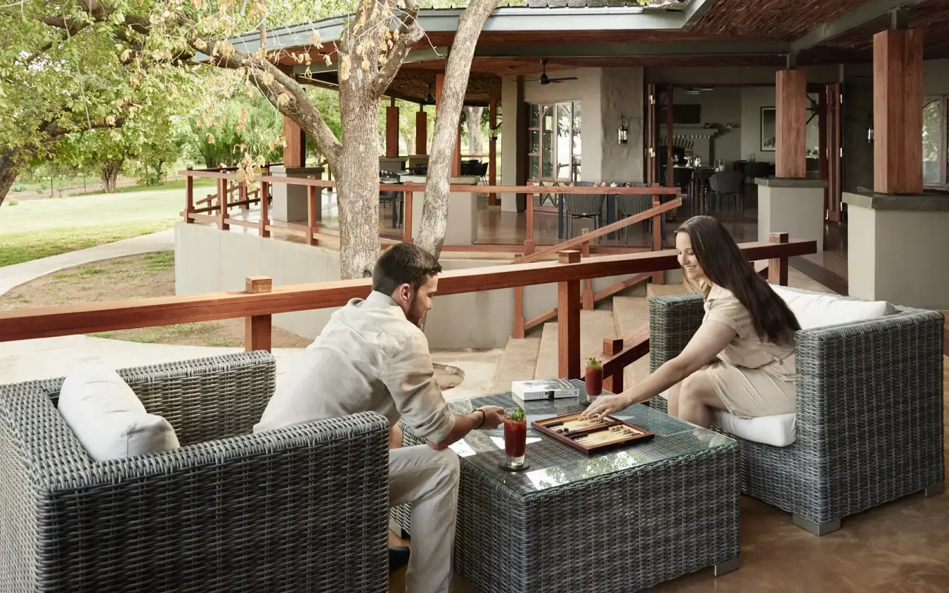 Couple relaxing on wicker furniture on shaded lodge veranda with trees, Chobe Chilwero, Chobe, Botswana